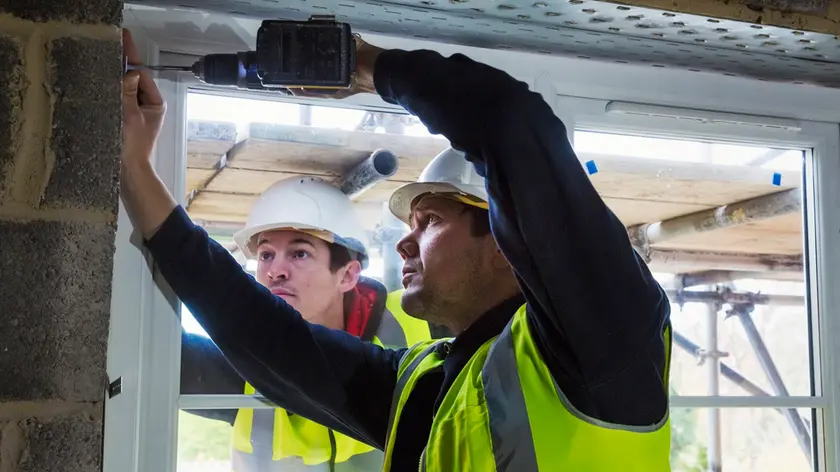 Two workmen on a construction site, builder in hard hat using an electric drill on a window frame.