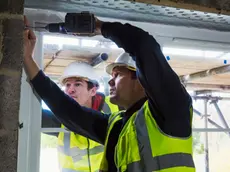 Two workmen on a construction site, builder in hard hat using an electric drill on a window frame.