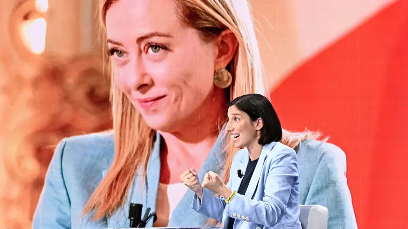 Nella foto Elly Schlein, sullo sfondo un’immagine della Meloni