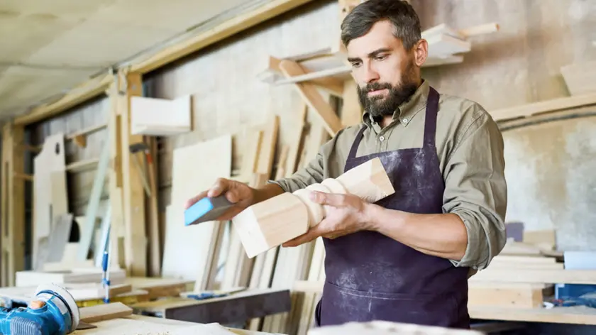 Handsome bearded craftsman using sandpaper in order to remove paint from wooden detail, interior of spacious workshop on background