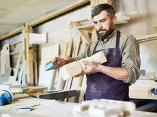 Handsome bearded craftsman using sandpaper in order to remove paint from wooden detail, interior of spacious workshop on background