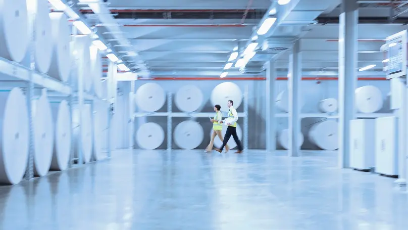 Workers walking along large paper spools in printing plant